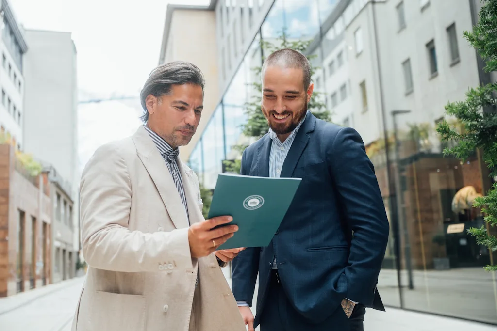 Zwei Geschäftsmänner im Anzug besprechen Unterlagen vor modernem Gebäude – Internationale Promotionsberatung